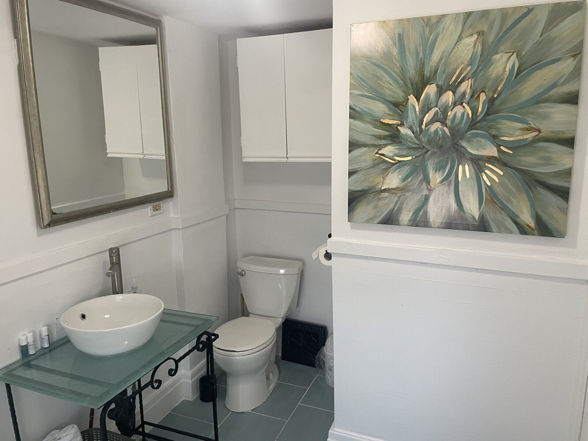 A bathroom with a glass sink, white toilet, and a large floral painting on the wall.