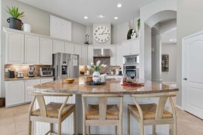 Una cocina moderna con armarios blancos, una isla central con taburetes de bar, un reloj de pared grande y varios electrodomésticos.