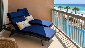Varanda com duas cadeiras de descanso azuis e uma cadeira de vime, com vista para uma piscina e uma praia.