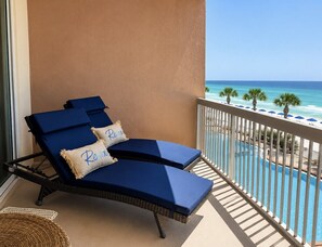 A balcony with two blue lounge chairs and a wicker chair, overlooking a pool and beach.