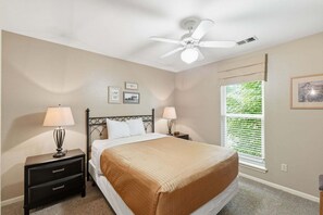A bedroom with a bed, two bedside tables with lamps, a ceiling fan, a window with blinds, and framed pictures on the wall.