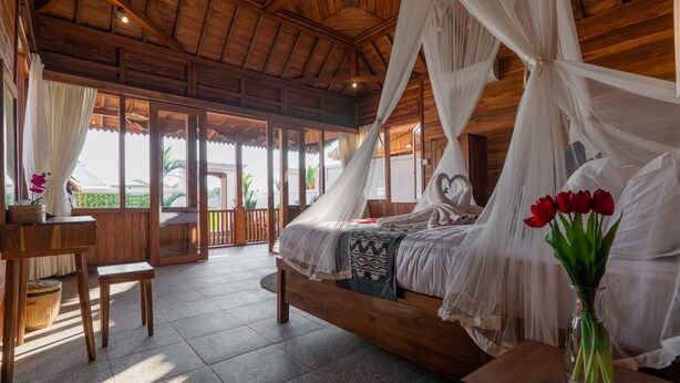 A wooden room with a bed under a canopy, a table with a vase of flowers, and a view of greenery outside. - Pondok di Uma Villa (Tegallalang)