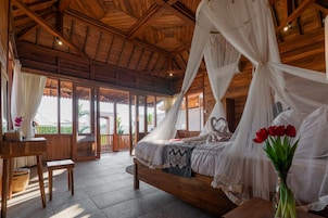 Una habitación de madera con una cama bajo un dosel, una mesa con un florero y vistas a la vegetación exterior.