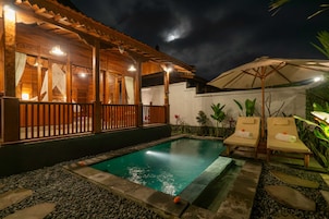 Un área de piscina nocturna con terraza de madera, sillones de descanso y una piscina pequeña.