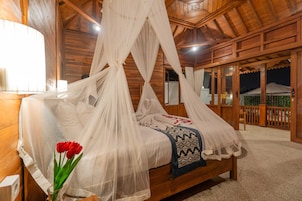 Una habitación de madera con una cama cubierta por una mosquitera blanca, un florero con flores rojas y una manta estampada.