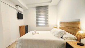 Ein ordentlich eingerichtetes Schlafzimmer mit einem Bett, einem Nachttisch aus Holz, einer Lampe, einem Fernseher und einem Fenster mit Jalousien.