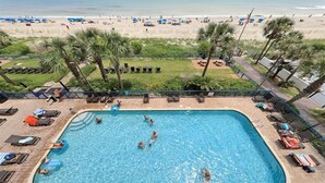 Ein Schwimmbad mit Schwimmern und Liegebädern, ein Strand mit Sonnenschirmen und Personen im Hintergrund sowie Palmen.