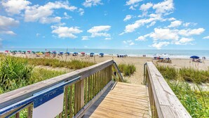 A wooden boardwalk leading to a beach with umbrellas and people.
