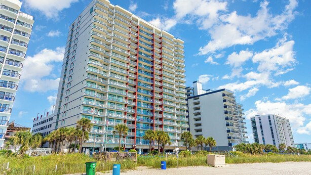 A multi-story hotel with balconies, located near a beach with palm trees and a sandy area.