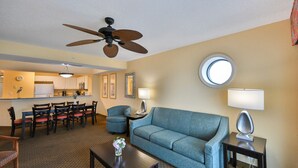 A hotel room with a dining area, a sofa, a coffee table, a lamp, a ceiling fan, and a window.