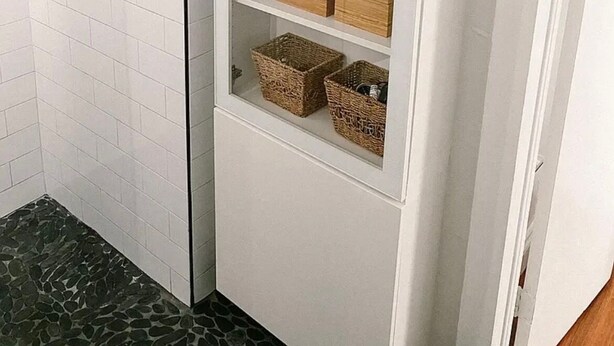 Een witte opslagunit met planken, gevuld met toiletpapier, houten dozen en rieten manden.