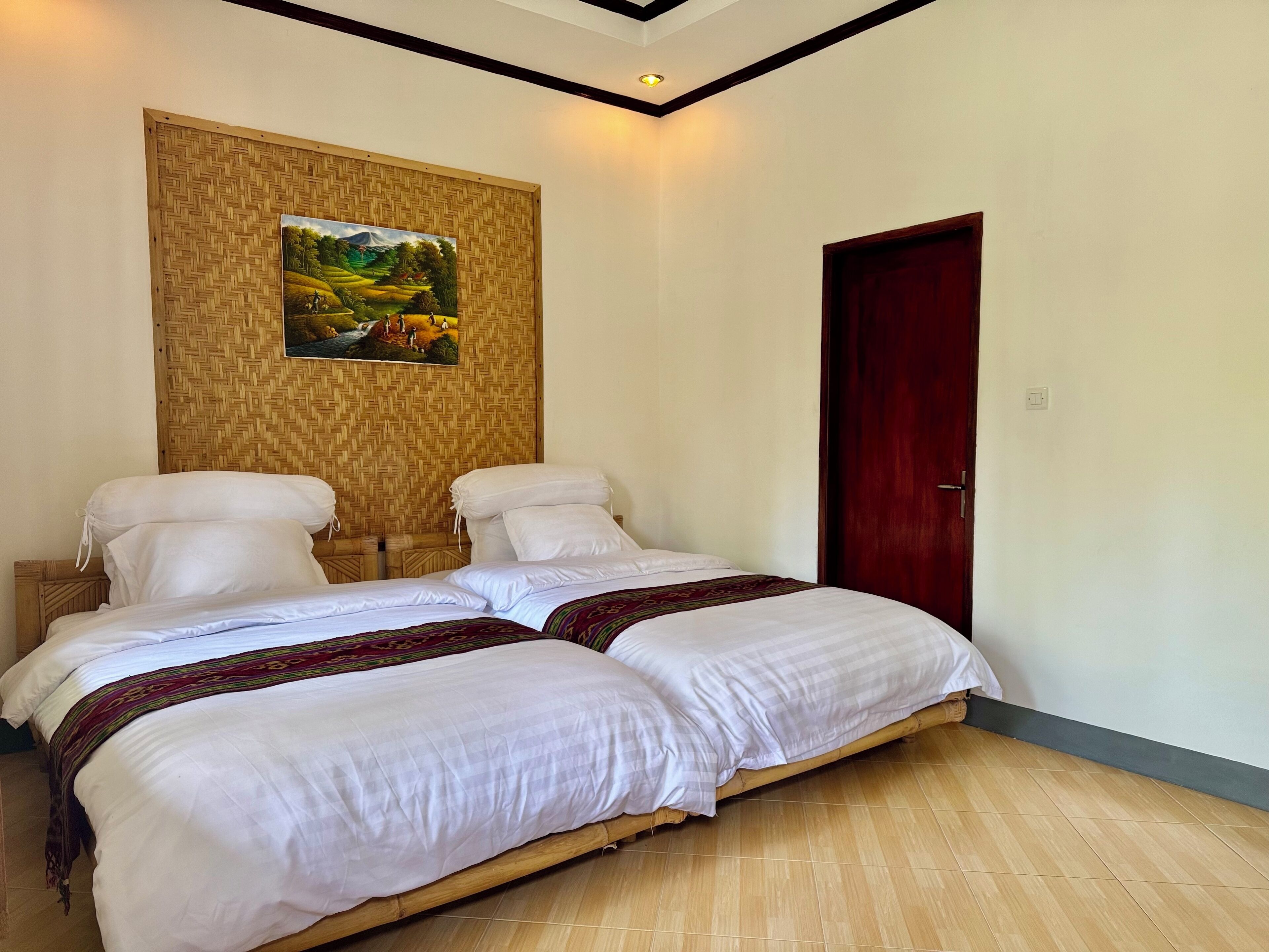 A double bed with white linens and a decorative throw blanket, a wooden headboard, and a woven wall panel with a landscape painting.