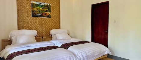 A double bed with white linens and a decorative throw blanket, a wooden headboard, and a woven wall panel with a landscape painting.