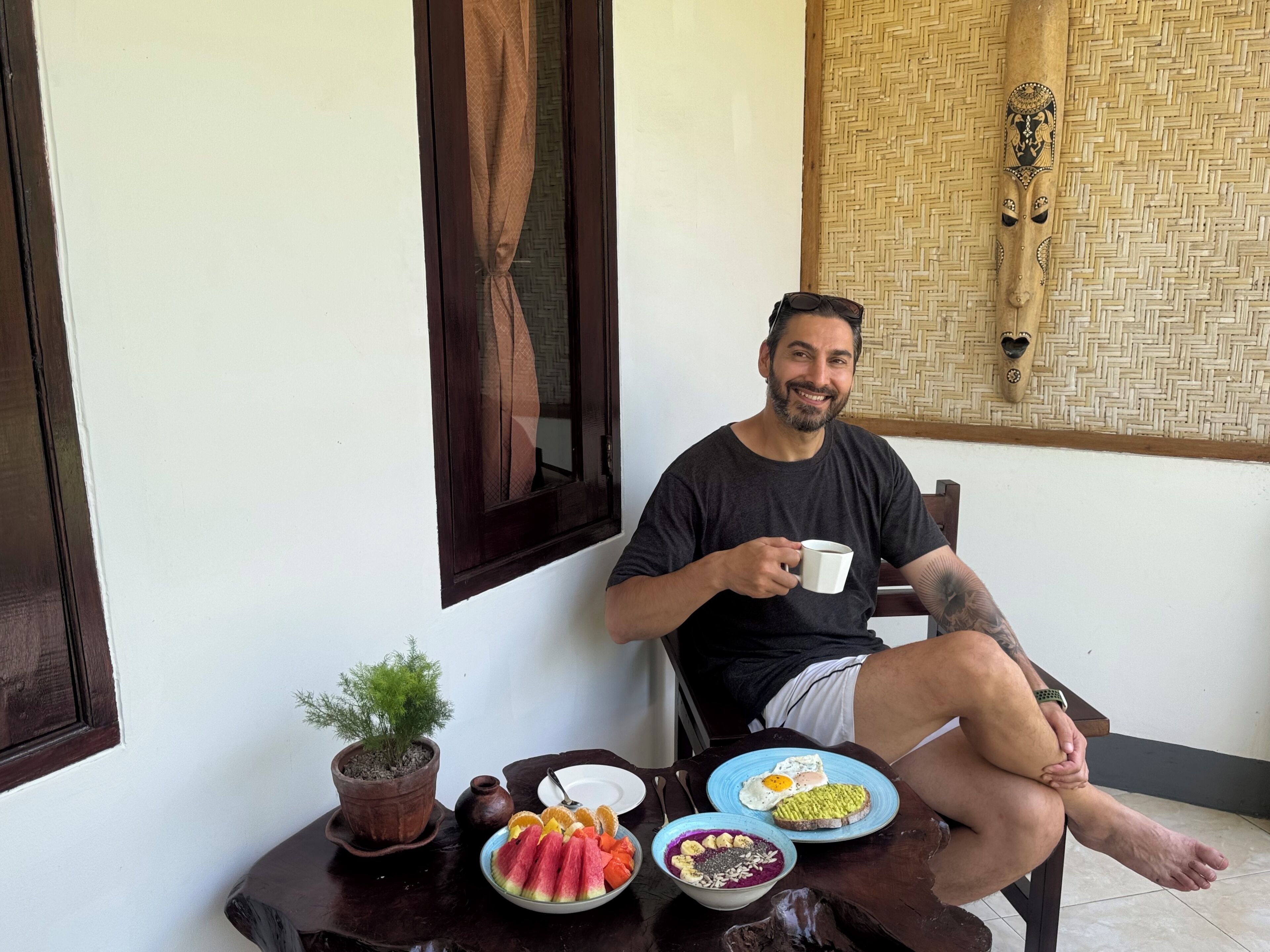 A man sitting on a wooden chair, holding a cup, with a table in front of him containing plates of food, a potted plant, and a woven wall hanging.