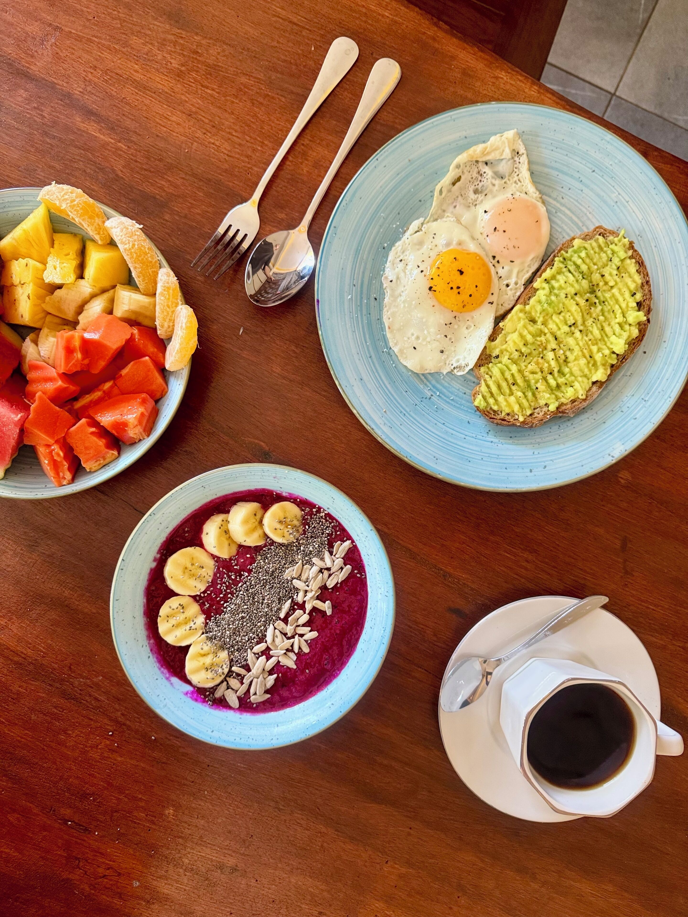 A breakfast table with a plate of scrambled eggs, toast with avocado, a bowl of fruit, and a bowl of smoothie.