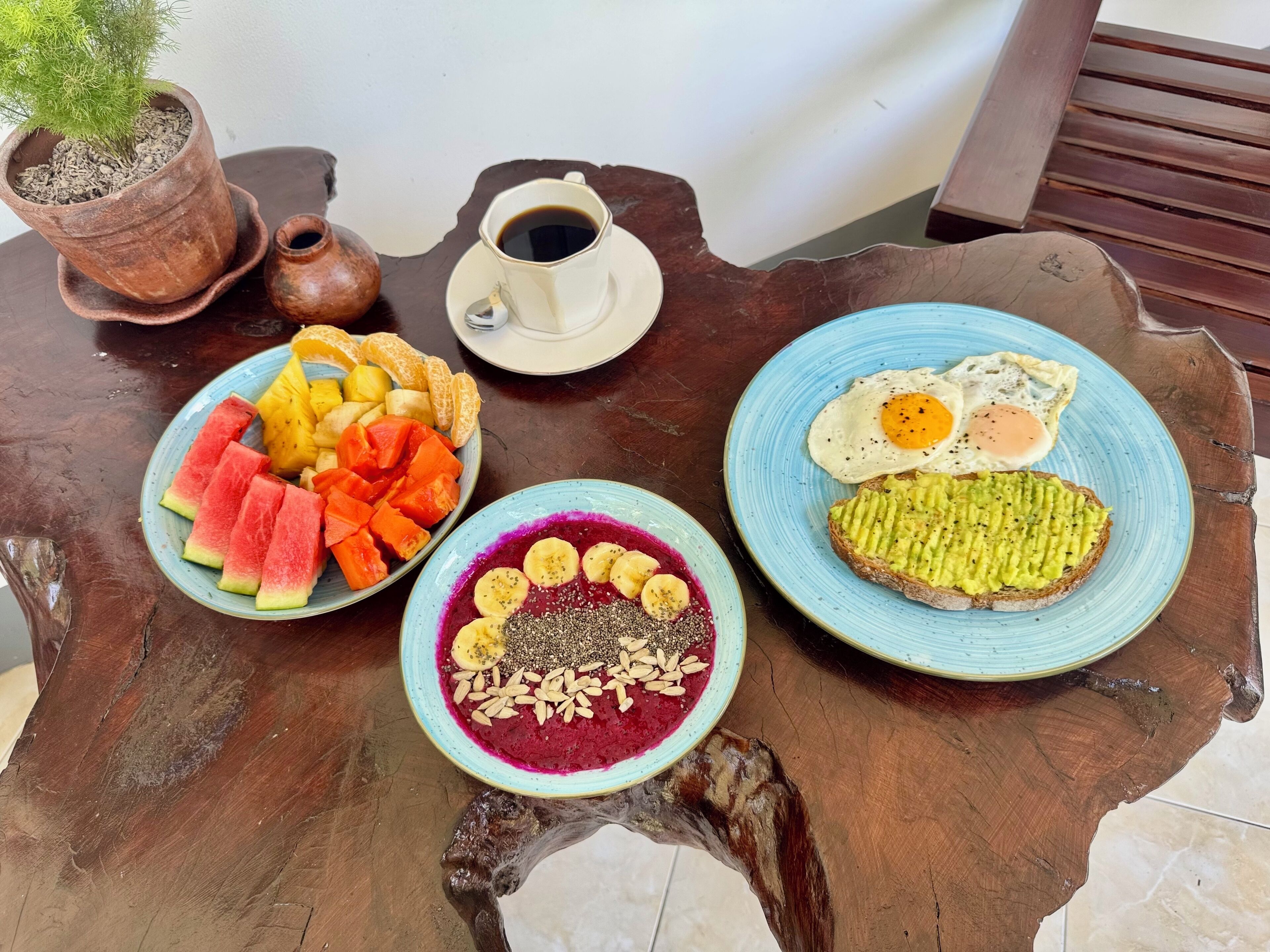 A wooden table with a plate of fruit, a bowl of smoothie, and a plate with eggs and toast.