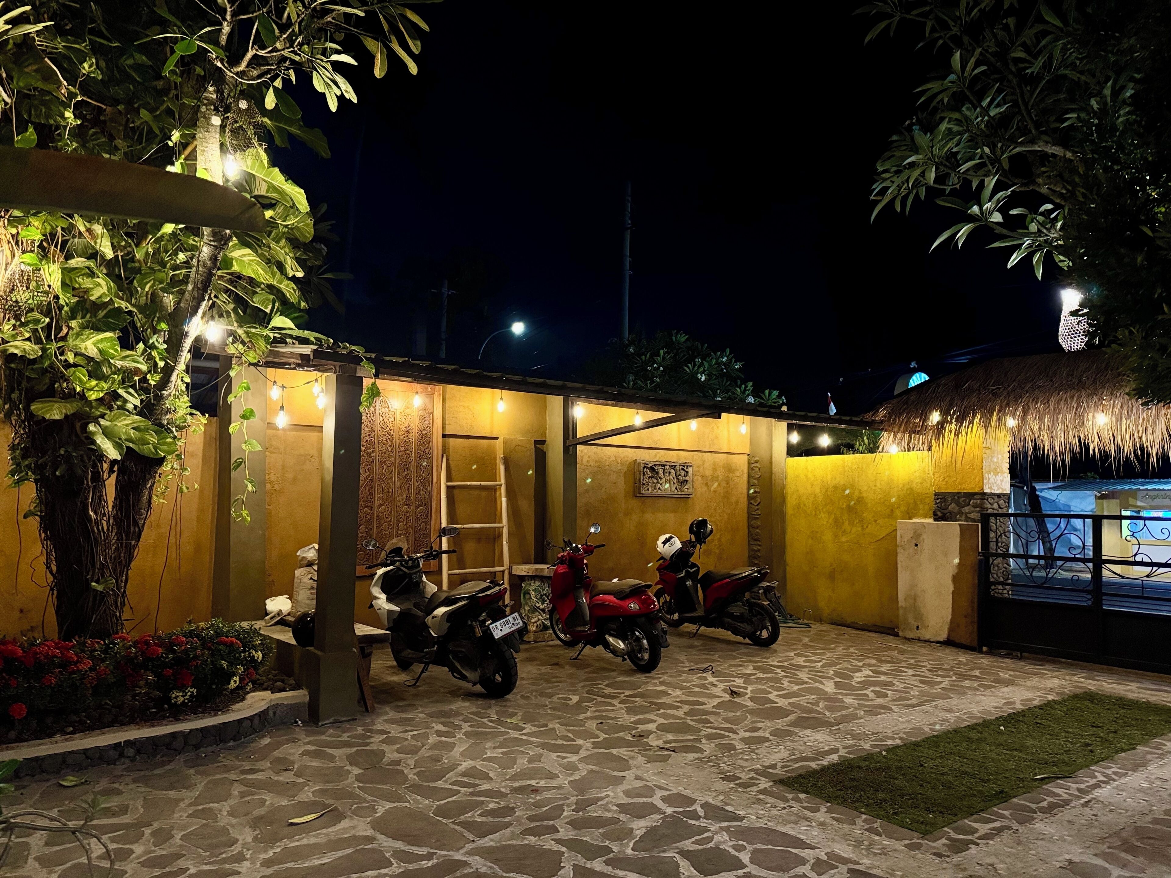 A nighttime outdoor area with a stone-paved patio, two motorcycles parked, and a thatched-roof structure.