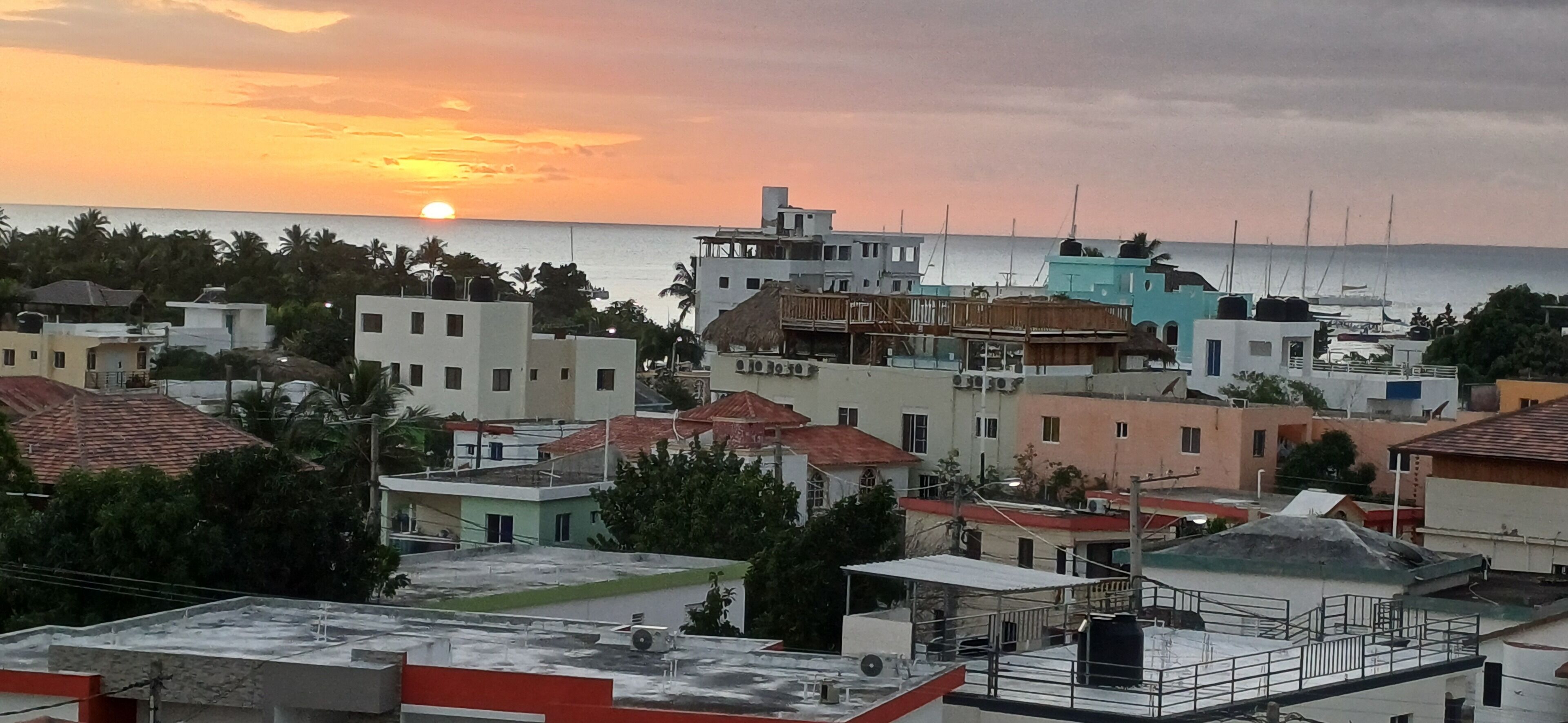 Una cittadina costiera con edifici, alcuni con tetti rossi, e un tramonto sul mare.