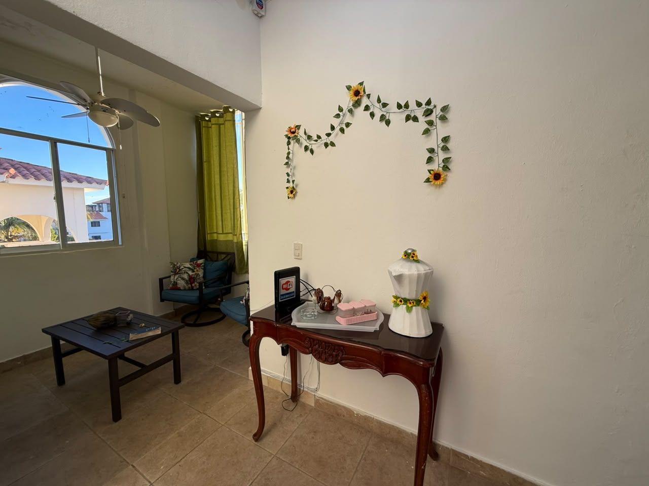 A room with a ceiling fan, a window with a view, a table with a decorative item, and a wall with a floral arrangement.