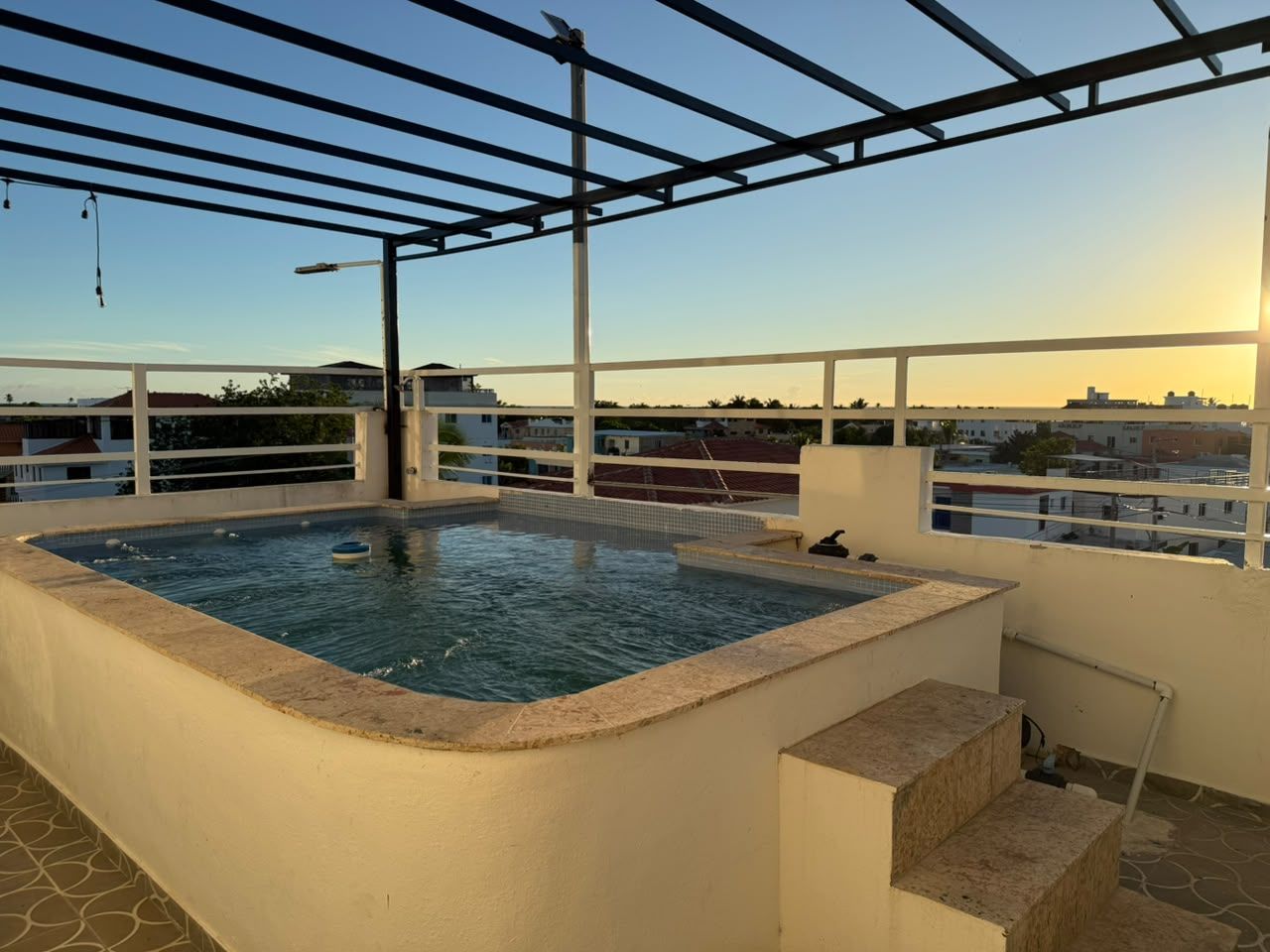 A rooftop swimming pool with a pergola structure, surrounded by a white wall and a clear view of the cityscape.