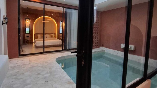 A modern hotel room with a glass door leading to a bedroom, a swimming pool, and a ceiling with circular light fixtures.