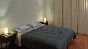 Een eenpersoonskamer met een houten vloer, twee nachtkastjes met lampen, een donkerdekkbed en een raam met verticale jaloezieën.