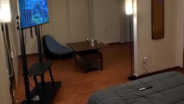 Een hotelkamer met een bed, een tv op een standaard, een stoel, een klein tafeltje en een deur naar een andere kamer.