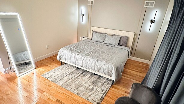A modern bedroom with a large bed, a bedside table, a mirror, and a chair. - Luxurious 3-bedroom 2 bath near NYC train station, EWR. (Newark)
