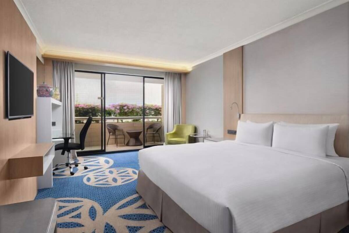 A hotel room with a bed, a desk, a chair, a TV, and a balcony with a view of flowers.