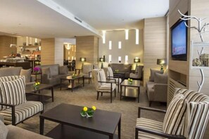 Un vestíbulo de hotel con una zona de estar, un televisor montado en la pared y una cocina visible al fondo.