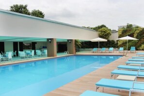 Une piscine entourée de chaises longues et de parasols, elle-même bordée d’une zone couverte avec des tables et des chaises.