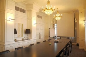 Een vergaderruimte met een lange tafel, stoelen, een kroonluchter, een whiteboard en een projectiescherm.