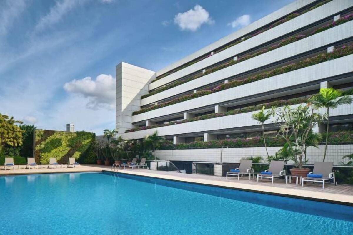 A modern hotel with a pool, multiple balconies with plants, and a clear sky.