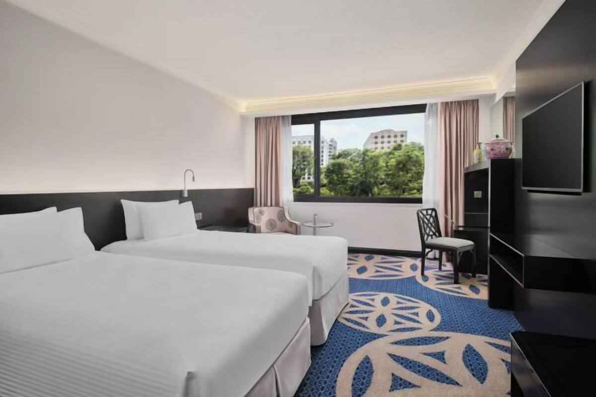 A hotel room with two beds, a TV, a desk, and a view of greenery outside.