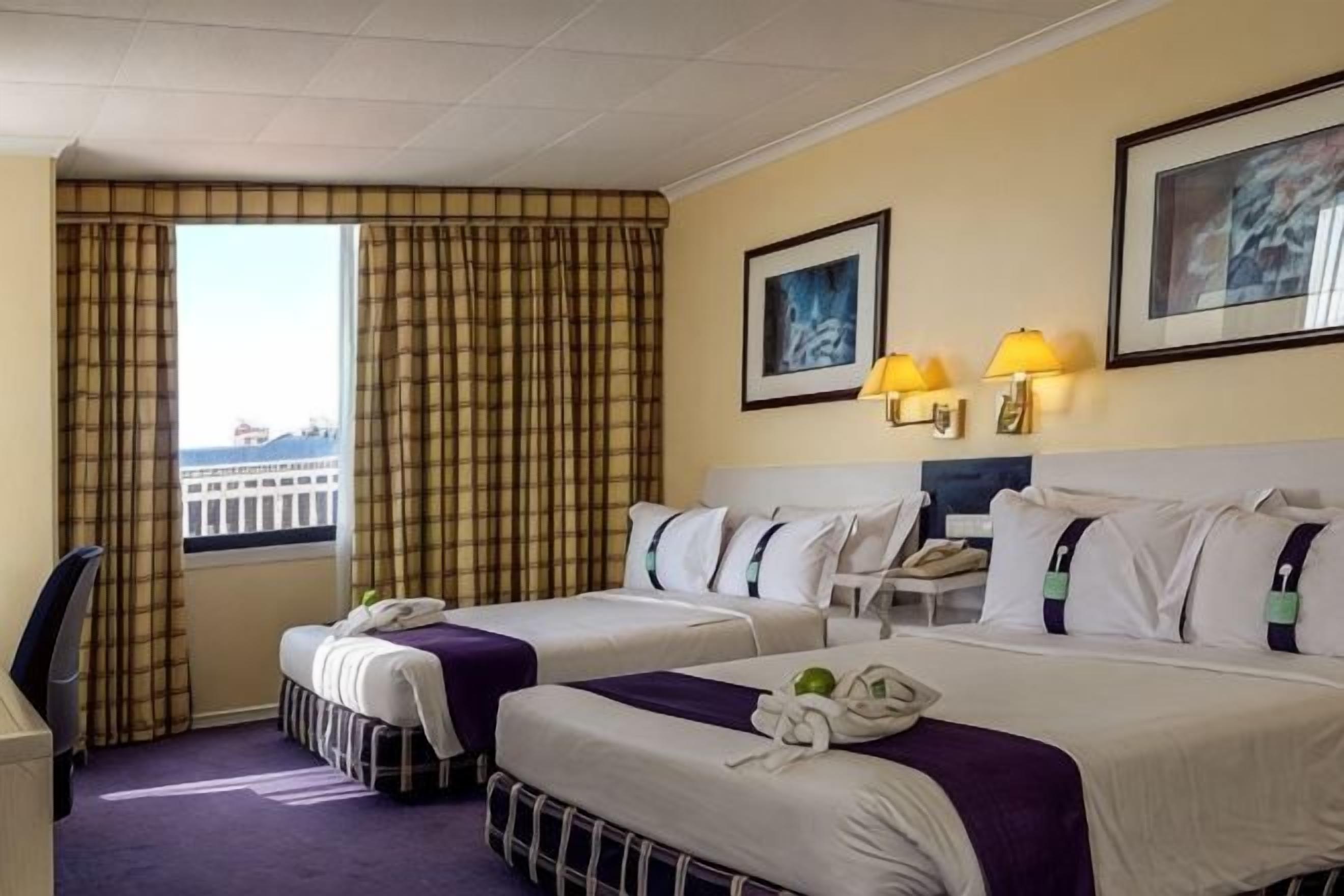 Une chambre d’hôtel avec deux lits, un bureau, une chaise, une fenêtre avec des rideaux et deux lampes fixées au mur.