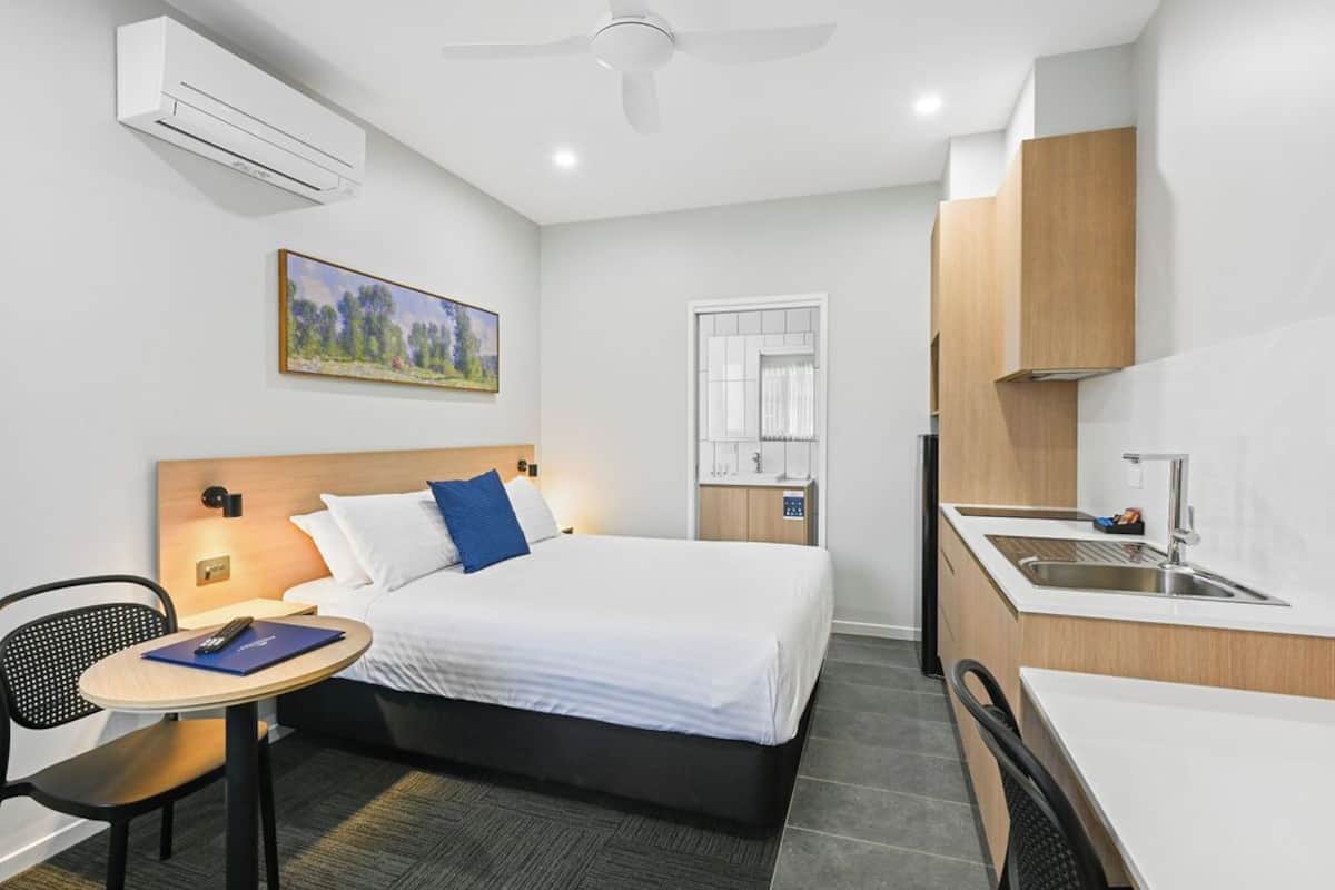 A hotel room with a bed, a desk, a chair, a small table, a painting, an air conditioner, a ceiling fan, a window, and a sink.