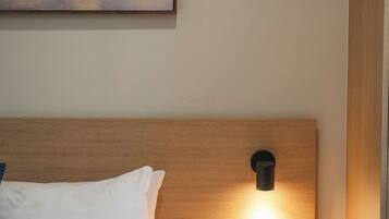 Una habitación de hotel con una cama, una mesita de noche con una lámpara fijada a la pared, y un cuadro sobre la cama.