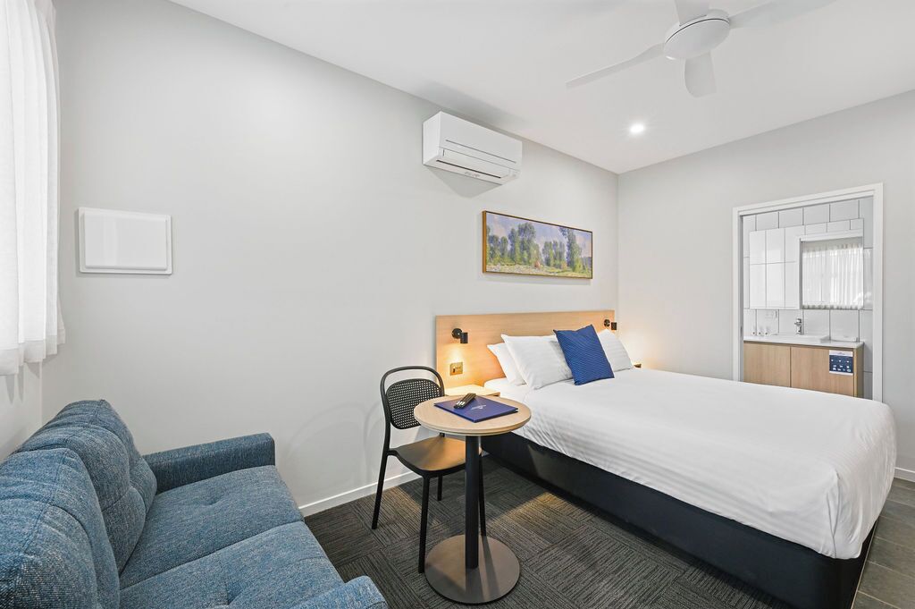 Habitación de hotel con cama, escritorio con silla, sofá, equipo de aire acondicionado, un cuadro en la pared y ventana con cortinas.