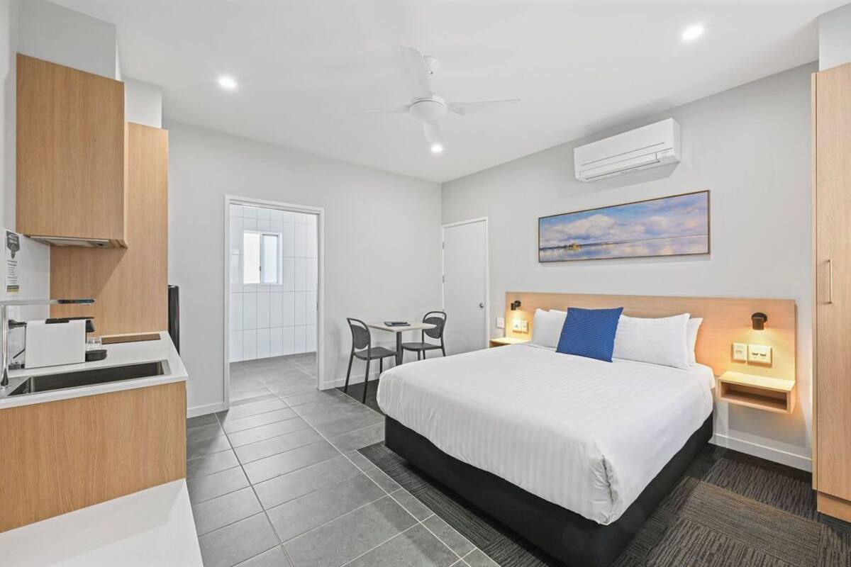 A hotel room with a bed, bedside tables, a desk with a chair, a ceiling fan, an air conditioning unit, and a painting on the wall.