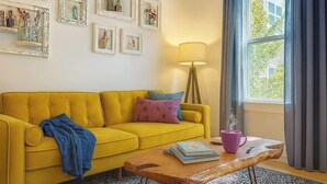 Un canapé jaune, une table basse en bois, une lampe et des œuvres d’art encadrées accrochées au mur.