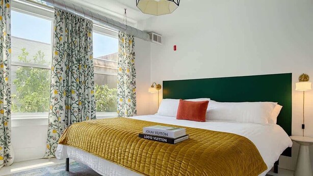 Une chambre d’hôtel avec un lit, une tête de lit verte, une fenêtre avec des rideaux et une table de chevet avec une lampe.