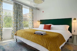 Quarto de hotel com cama, cabeceira verde, janela com cortinas e mesa de cabeceira com abajur.