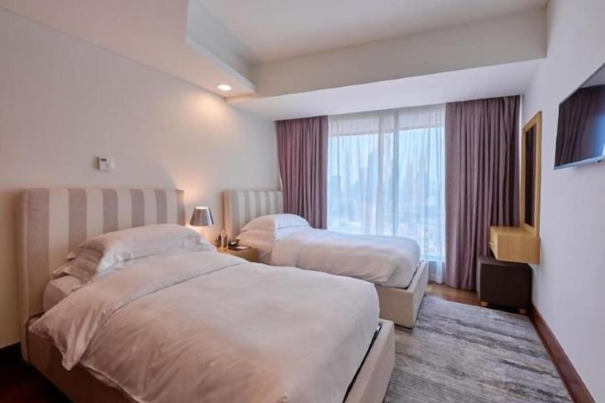 Ein Hotelzimmer mit zwei Betten, einem großen Fenster mit Vorhängen, einem Flachbildfernseher und einem kleinen Tisch.