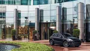 Een modern gebouw met een glazen gevel, een zwarte auto geparkeerd ervoor, en een aangelegde tuin met een fontein.
