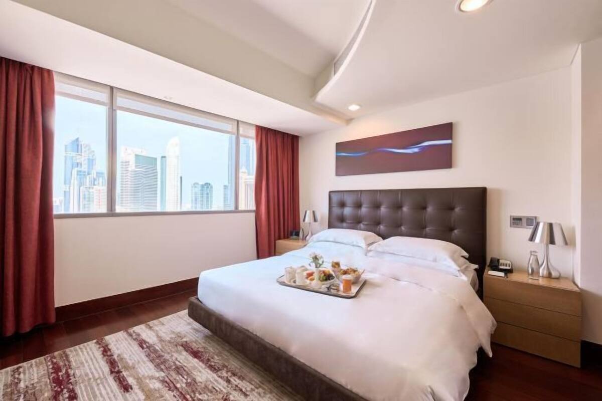A hotel room with a large bed, a city view, a modern headboard, and a tray of breakfast items.
