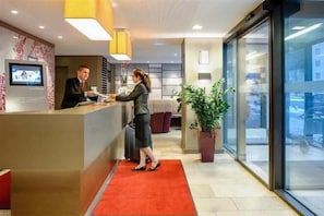 A reception desk with a man and a woman, a large potted plant, and a red carpet. - Mercure München City Schwabing (Munich)