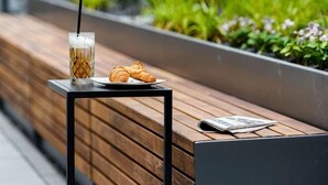 A wooden bench with a small table, a glass of iced coffee, a croissant, and a magazine.