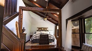 Un dormitorio con techo de madera, una cama con cabecera, una ventana con vistas a zonas verdes y un ventilador en el alféizar.