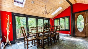 Una habitación con suelo de piedra, techo de madera y ventanas amplias. Se observan varias sillas y una mesa de madera.
