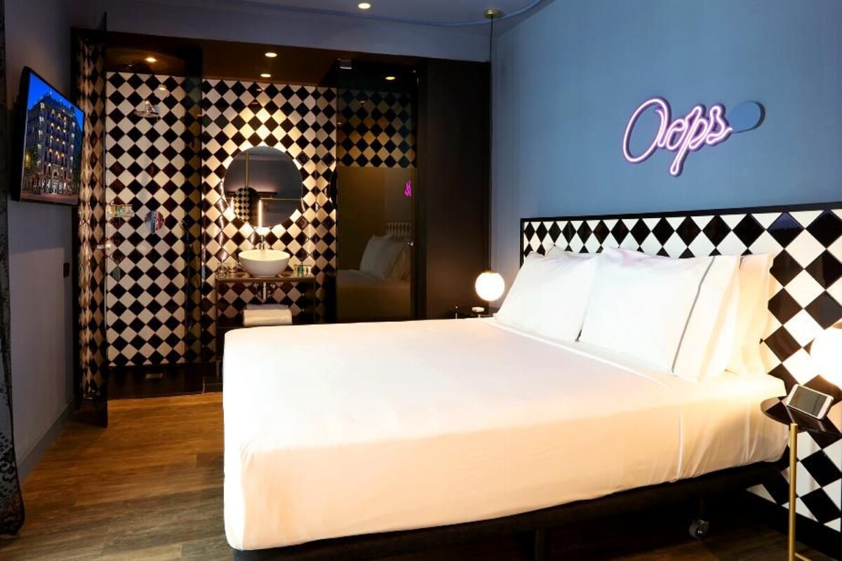 A hotel room with a bed, a bedside table with a lamp, a mirror, and a neon sign on the wall.